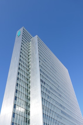 A tall, modern skyscraper with a grid-like facade of windows and a metallic exterior. The building rises against a clear blue sky, with a logo displayed near its rooftop.