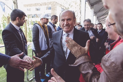 A group of people gathered outdoors, with a central figure in a suit smiling and shaking hands with several individuals. The setting appears to be a formal or public event, with a mix of casual and dressed-up attendees.