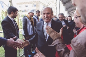 A group of people gathered outdoors, with a central figure in a suit smiling and shaking hands with several individuals. The setting appears to be a formal or public event, with a mix of casual and dressed-up attendees.