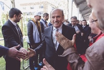 A group of people gathered outdoors, with a central figure in a suit smiling and shaking hands with several individuals. The setting appears to be a formal or public event, with a mix of casual and dressed-up attendees.