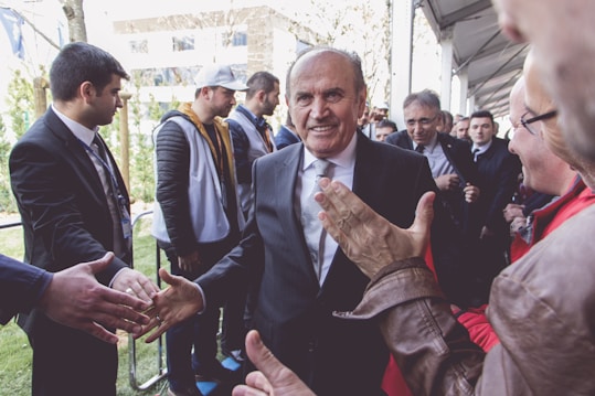 A group of people gathered outdoors, with a central figure in a suit smiling and shaking hands with several individuals. The setting appears to be a formal or public event, with a mix of casual and dressed-up attendees.