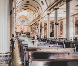 An ornate interior of a church with tall columns, wooden pews, and a decorated altar with religious artwork in the background. The architecture is traditional with arched ceilings and intricate details. A few people are scattered throughout the pews, creating a sense of tranquility and reflection.