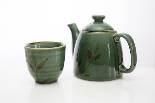 A green ceramic teapot with a matching cup sits on a smooth white surface. Both the teapot and cup feature a subtle leaf pattern, adding a touch of nature-inspired design to their appearance.