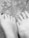 Two feet are placed side by side on a gritty surface, possibly sand. One foot has nail polish on some toes, indicating a possible casual setting. The texture of the surface contrasts with the smoothness of the feet.