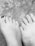 Two feet are placed side by side on a gritty surface, possibly sand. One foot has nail polish on some toes, indicating a possible casual setting. The texture of the surface contrasts with the smoothness of the feet.