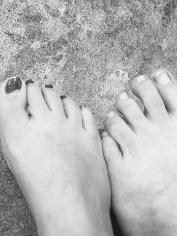 Two feet are placed side by side on a gritty surface, possibly sand. One foot has nail polish on some toes, indicating a possible casual setting. The texture of the surface contrasts with the smoothness of the feet.