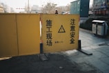 A yellow construction barrier with warning signs in Chinese. The barrier is placed outdoors on a paved area near some vegetation and utility boxes. A person is partially visible behind the barrier.