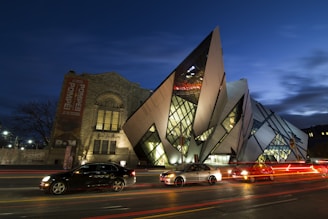 An artistic, modern building with sharp, angular designs and glass panels is prominently featured. To the left, there is an older, more traditional stone building with a large vertical banner advertising an exhibit. Several cars with light trails from the street are visible in the foreground, suggesting a busy urban setting at night.