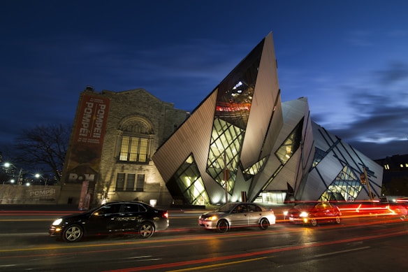 An artistic, modern building with sharp, angular designs and glass panels is prominently featured. To the left, there is an older, more traditional stone building with a large vertical banner advertising an exhibit. Several cars with light trails from the street are visible in the foreground, suggesting a busy urban setting at night.