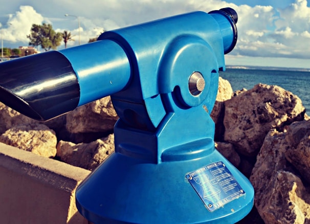 A blue public viewing telescope positioned on a rocky seaside location. The sea is visible in the background under a partly cloudy sky, adding an element of natural beauty. The telescope appears to be fixed on a pedestal, with rocks surrounding the area, and distant trees and a road visible.