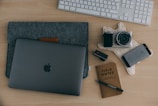 A neatly arranged workspace on a wooden table, including a laptop in a grey sleeve, a digital camera on a textured cloth, a smartphone, a USB stick, a Field Notes notebook with a pen, and a wireless keyboard in the background.
