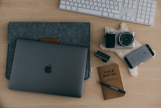 A neatly arranged workspace on a wooden table, including a laptop in a grey sleeve, a digital camera on a textured cloth, a smartphone, a USB stick, a Field Notes notebook with a pen, and a wireless keyboard in the background.