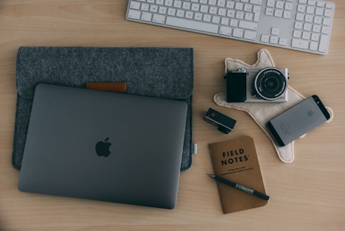 A neatly arranged workspace on a wooden table, including a laptop in a grey sleeve, a digital camera on a textured cloth, a smartphone, a USB stick, a Field Notes notebook with a pen, and a wireless keyboard in the background.