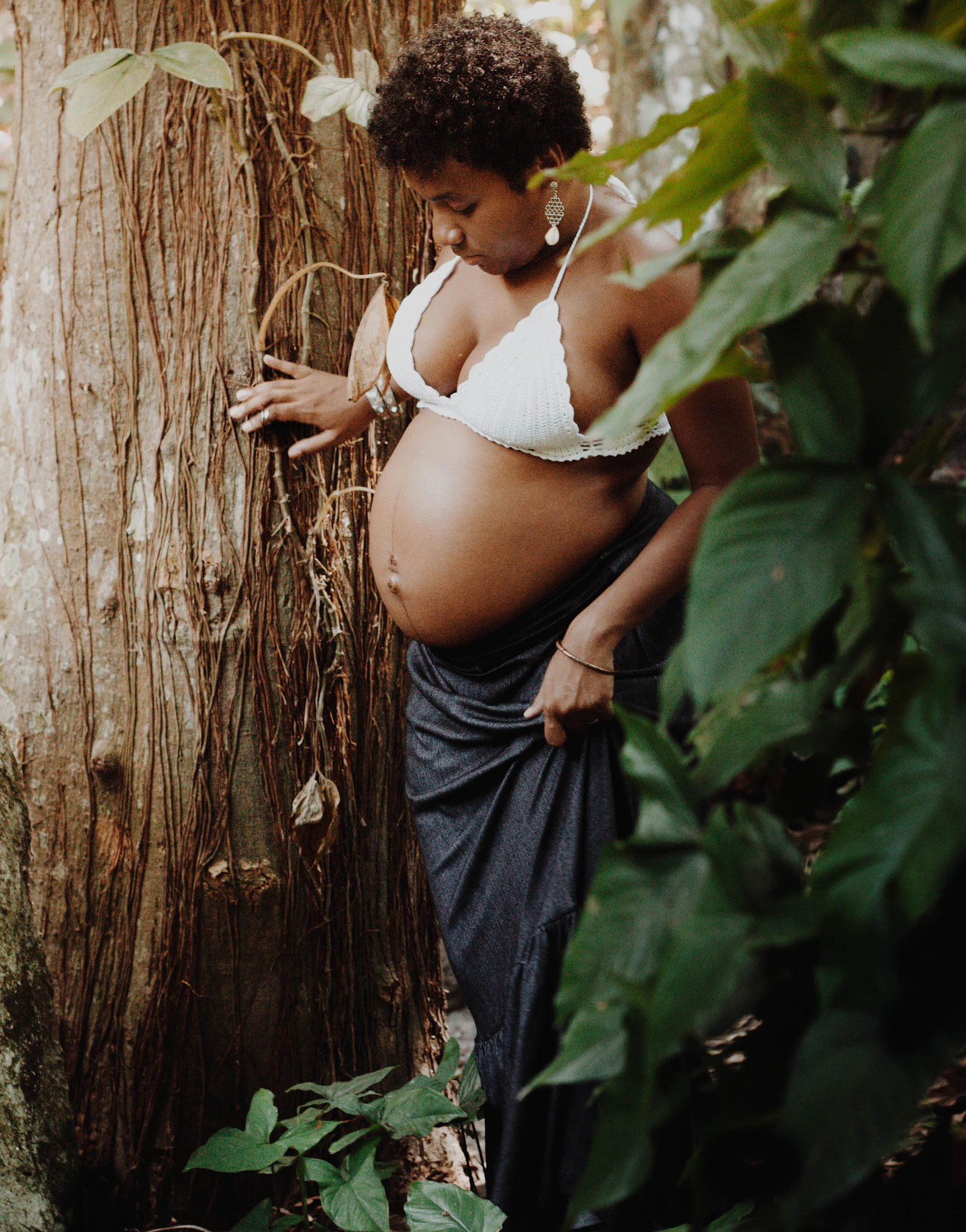 A pregnant woman in a light dress leaning against a tree trunk in a lush green forest.