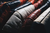 A close-up view of several leather jackets hanging on a rack, showcasing different colors such as black, red, white, and brown. The texture of the leather is highlighted, with visible stitching and wear.