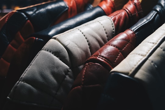 A close-up view of several leather jackets hanging on a rack, showcasing different colors such as black, red, white, and brown. The texture of the leather is highlighted, with visible stitching and wear.