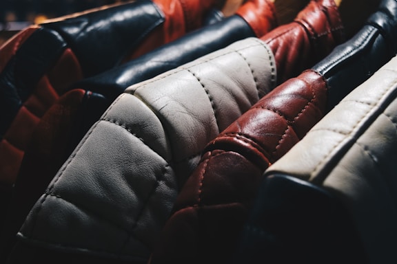 A close-up view of several leather jackets hanging on a rack, showcasing different colors such as black, red, white, and brown. The texture of the leather is highlighted, with visible stitching and wear.