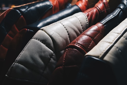 A close-up view of several leather jackets hanging on a rack, showcasing different colors such as black, red, white, and brown. The texture of the leather is highlighted, with visible stitching and wear.