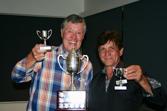 Two men are smiling and holding trophies, with one wearing a colorful plaid shirt and the other in a dark patterned shirt. The larger trophy is held centrally, while each man holds a smaller one.