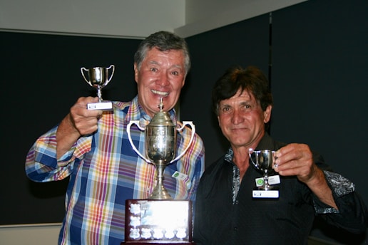 Two men are smiling and holding trophies, with one wearing a colorful plaid shirt and the other in a dark patterned shirt. The larger trophy is held centrally, while each man holds a smaller one.