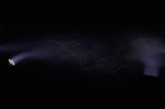Two beams of light punctuate the darkness, casting subtle glows across the scene. The left beam emanates from a light source with a prism effect, projecting a small rainbow onto the darkness. Both beams fade softly into the background, creating an ethereal ambiance.