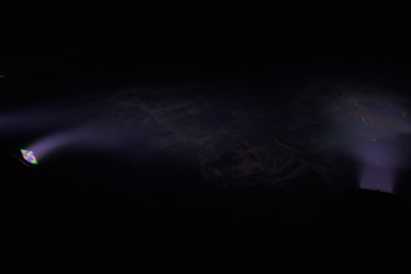 Two beams of light punctuate the darkness, casting subtle glows across the scene. The left beam emanates from a light source with a prism effect, projecting a small rainbow onto the darkness. Both beams fade softly into the background, creating an ethereal ambiance.