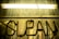 A misty glass panel with the word 'CLEAN' written in thick dirt or dust, set against a background illuminated by a glowing yellow fluorescent light. The surface of the glass appears weathered and grimy, suggesting a need for cleaning or neglect over time.