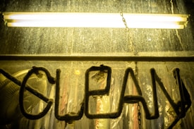 A misty glass panel with the word 'CLEAN' written in thick dirt or dust, set against a background illuminated by a glowing yellow fluorescent light. The surface of the glass appears weathered and grimy, suggesting a need for cleaning or neglect over time.