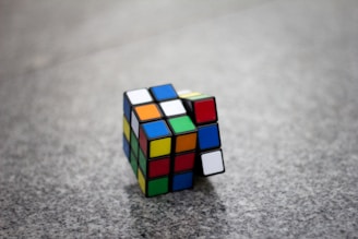 A partially solved Rubik's Cube rests on a speckled gray surface. The cube displays a mix of colors on its visible faces, with some rows aligned and others in disarray.