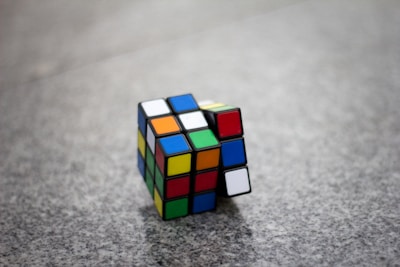 A partially solved Rubik's Cube rests on a speckled gray surface. The cube displays a mix of colors on its visible faces, with some rows aligned and others in disarray.