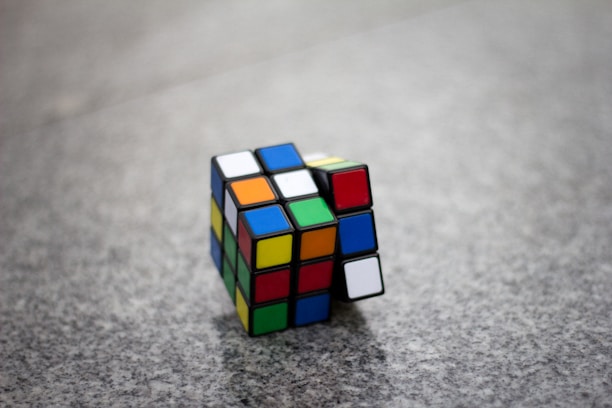 A partially solved Rubik's Cube rests on a speckled gray surface. The cube displays a mix of colors on its visible faces, with some rows aligned and others in disarray.