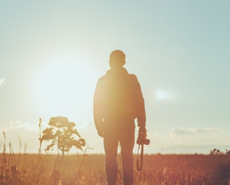 man standing on dried grass holding camera during golden hour