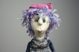 A whimsical doll with exaggerated facial features, including large round eyes and a prominent nose, is adorned with a vibrant pink bow on top of a head full of textured, colorful purple hair. The doll's expression is playful and slightly curious.