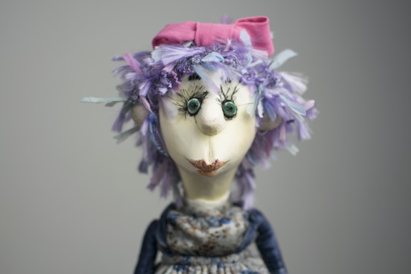 A whimsical doll with exaggerated facial features, including large round eyes and a prominent nose, is adorned with a vibrant pink bow on top of a head full of textured, colorful purple hair. The doll's expression is playful and slightly curious.
