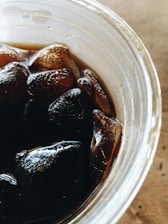 A close-up view of a transparent plastic cup filled with a dark liquid and several ice cubes. The surface of the liquid reflects light, providing a glossy appearance, while the ice cubes appear partially submerged and somewhat transparent.