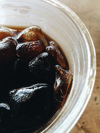 A close-up view of a transparent plastic cup filled with a dark liquid and several ice cubes. The surface of the liquid reflects light, providing a glossy appearance, while the ice cubes appear partially submerged and somewhat transparent.