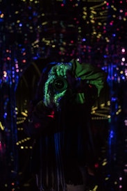 A person holding a camera is visible in the midst of a dark, reflective environment with multicolored lights. The scene is illuminated by small, scattered dots of blue, green, red, and yellow lights, creating a slightly surreal and abstract atmosphere. The reflective surfaces amplify the light patterns, giving a mirrored effect.