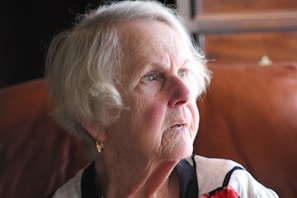 An elderly woman with short white hair is gazing thoughtfully, wearing a light blouse with dark accents. The lighting creates a warm ambience, highlighting her facial features and a pair of gold earrings.