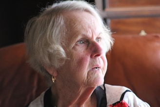 An elderly woman with short white hair is gazing thoughtfully, wearing a light blouse with dark accents. The lighting creates a warm ambience, highlighting her facial features and a pair of gold earrings.