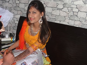 A woman with henna designs on her arm is sitting indoors, smiling at the camera. She is wearing traditional attire with prominent jewelry, including large earrings. The background features a wall with a stone pattern. There is a newspaper on her lap and someone else is applying henna to her hand.