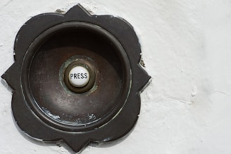 A vintage-style doorbell with a round metallic casing featuring a floral pattern around its edges. The center button is white with the word 'Press' written on it.