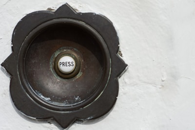 A vintage-style doorbell with a round metallic casing featuring a floral pattern around its edges. The center button is white with the word 'Press' written on it.