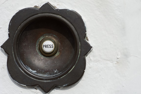 A vintage-style doorbell with a round metallic casing featuring a floral pattern around its edges. The center button is white with the word 'Press' written on it.