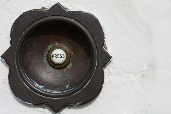 A vintage-style doorbell with a round metallic casing featuring a floral pattern around its edges. The center button is white with the word 'Press' written on it.