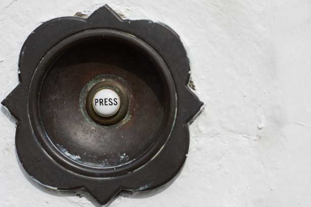 A vintage-style doorbell with a round metallic casing featuring a floral pattern around its edges. The center button is white with the word 'Press' written on it.