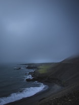 A dark, moody coastal landscape with rocky cliffs, a misty sky, and waves crashing against the shoreline. The terrain appears rugged and the atmosphere is overcast, conveying a sense of isolation and tranquility.