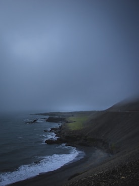 A dark, moody coastal landscape with rocky cliffs, a misty sky, and waves crashing against the shoreline. The terrain appears rugged and the atmosphere is overcast, conveying a sense of isolation and tranquility.