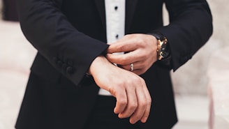 A person is adjusting the cuff of a tailored black suit jacket, wearing a white shirt underneath. The hands, adorned with a watch and a ring, are meticulously focused on the task, exuding a sense of elegance and sophistication.