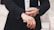 A person is adjusting the cuff of a tailored black suit jacket, wearing a white shirt underneath. The hands, adorned with a watch and a ring, are meticulously focused on the task, exuding a sense of elegance and sophistication.