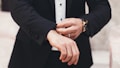 A person is adjusting the cuff of a tailored black suit jacket, wearing a white shirt underneath. The hands, adorned with a watch and a ring, are meticulously focused on the task, exuding a sense of elegance and sophistication.